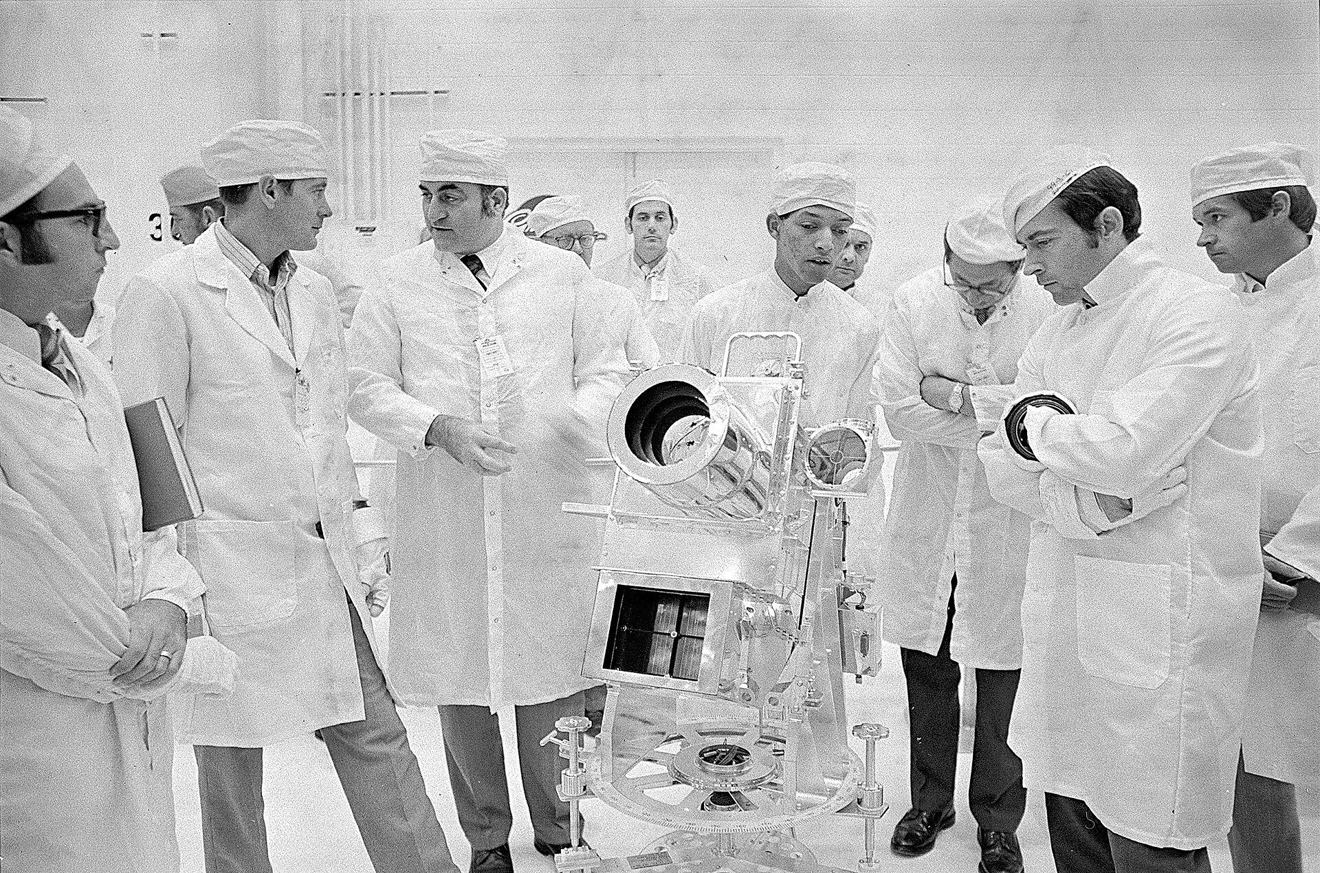 A black-and-white photo shows a group of at least 11 men wearing white lab coats and white caps; a telescopic object sits in front. Carruthers, in the center, is speaking.