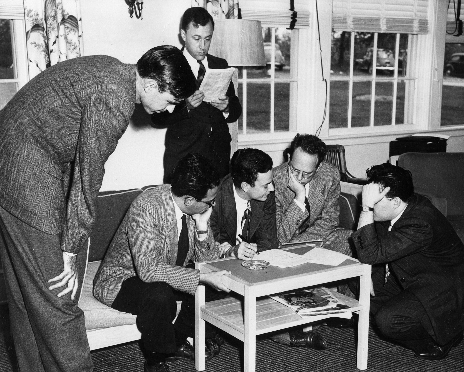 Willis Lamb, Abraham Pais, Richard Feynman, Herman Feshbach and Julian Schwinger dressed in suits with ties and gathered around a small table while deep in discussion during the Shelter Island Conference. 