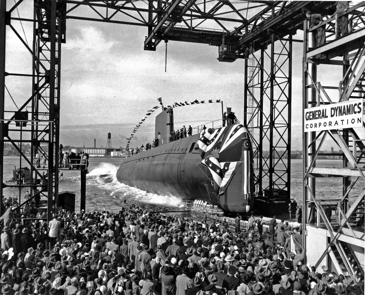 A large submarine launches from dock with huge crowd looking on.